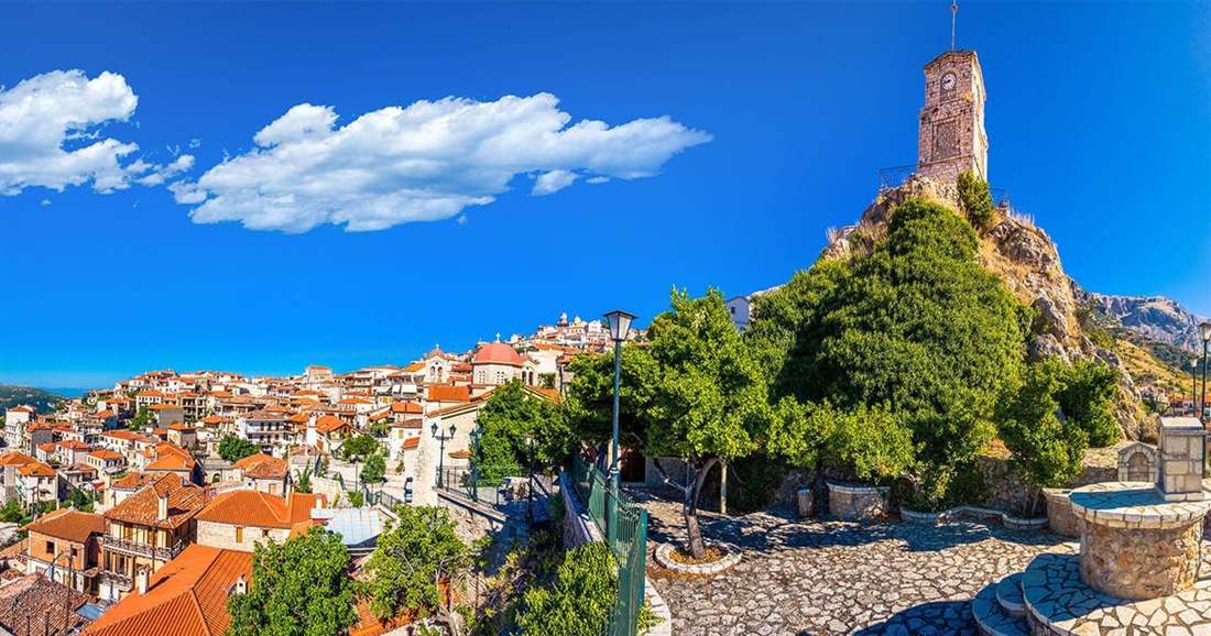 The clock tower of Arachova on a rock above red-tiled roofs, with the mountains of Parnassus under a blue sky.