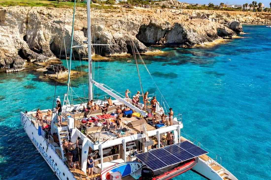 A catamaran full of guests anchored by the sea caves and cliffs of Cape Greco in turquoise water