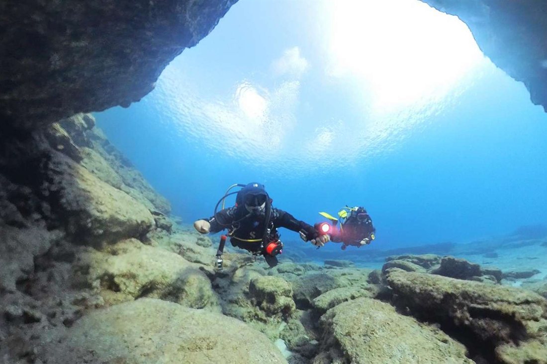 A scuba diver with a torch swims through a sunlit sea cave over a rocky seabed at Cape Greco
