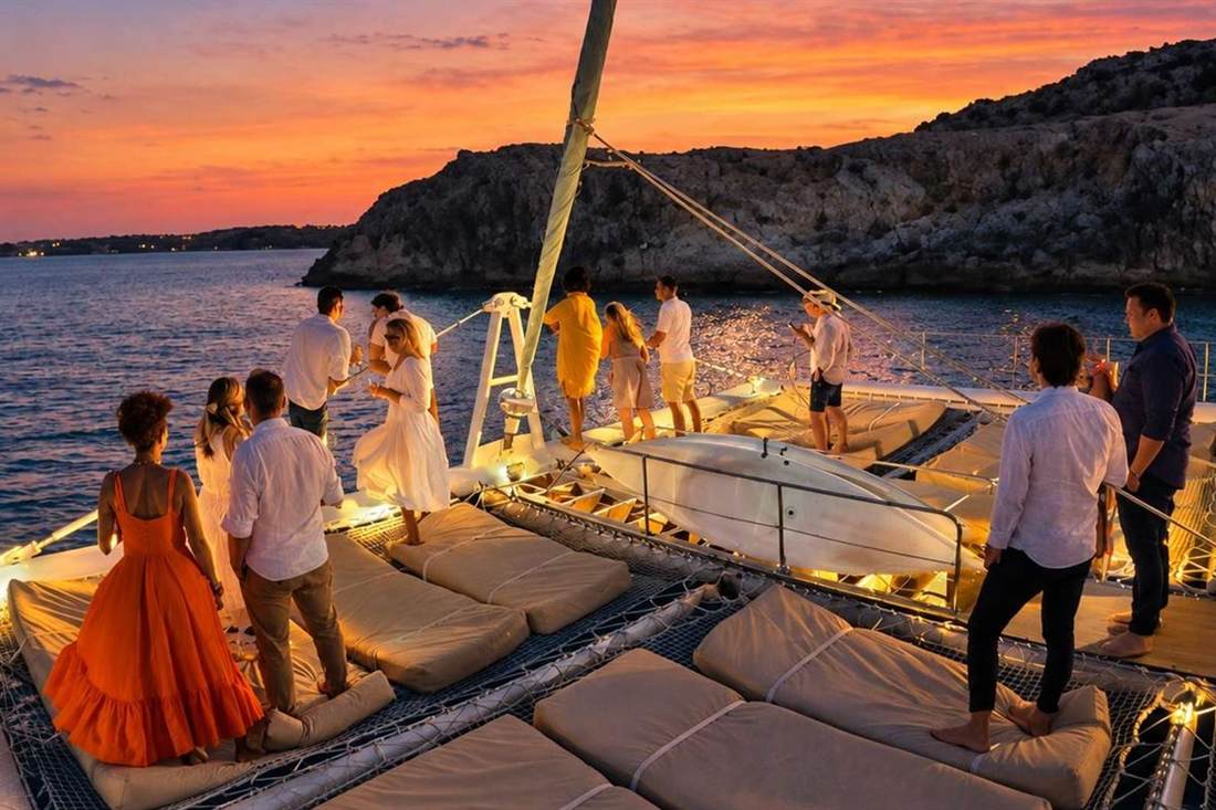 Guests in white and orange clothes stand on the net deck of a catamaran at sunset, with an orange sky over the sea and a rocky headland beside the boat