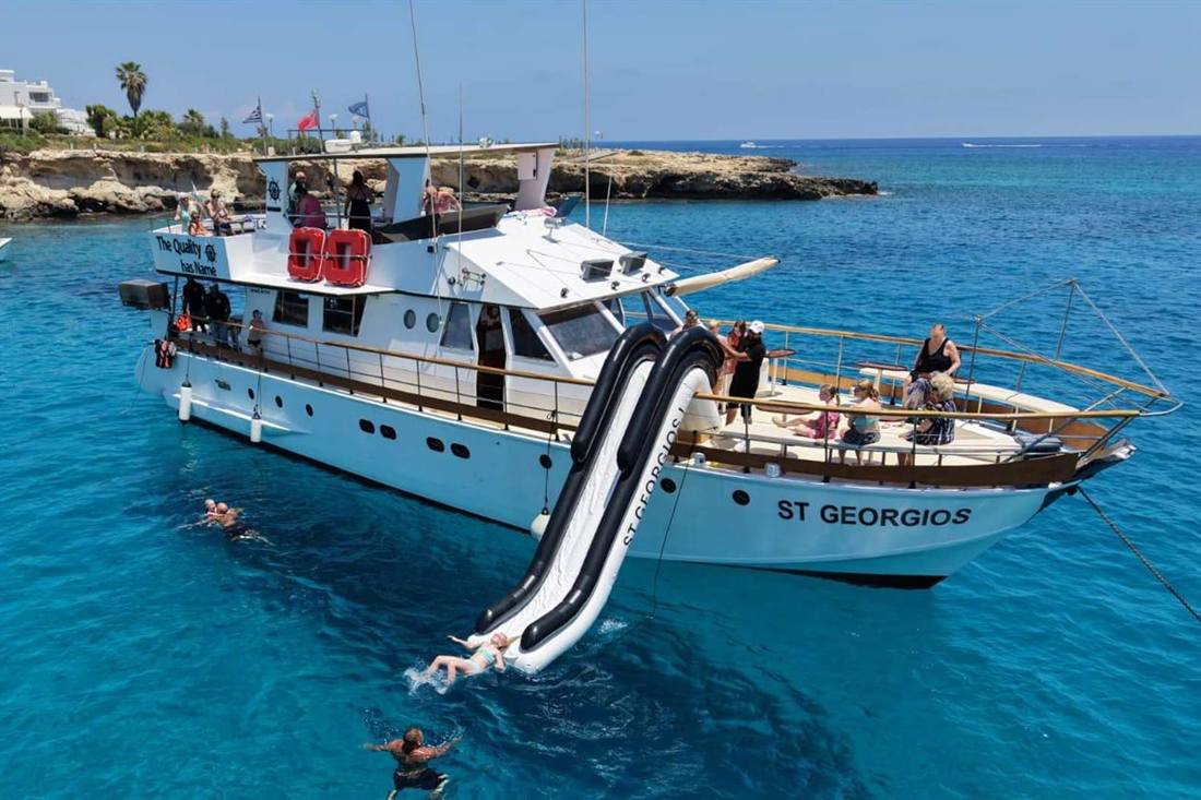 White cruise boat with a black and white water slide anchored in a turquoise cove, with swimmers in the water and a rocky shore behind it