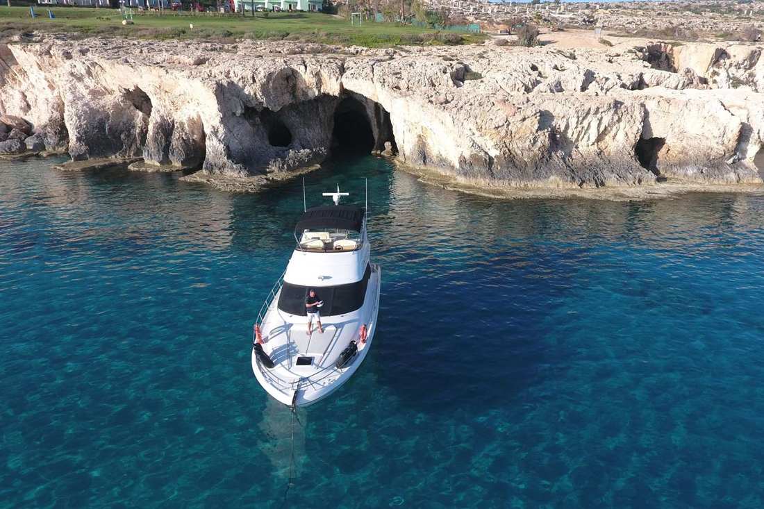 White motor yacht anchored in clear turquoise water in front of limestone cliffs with sea caves, a man standing on the bow
