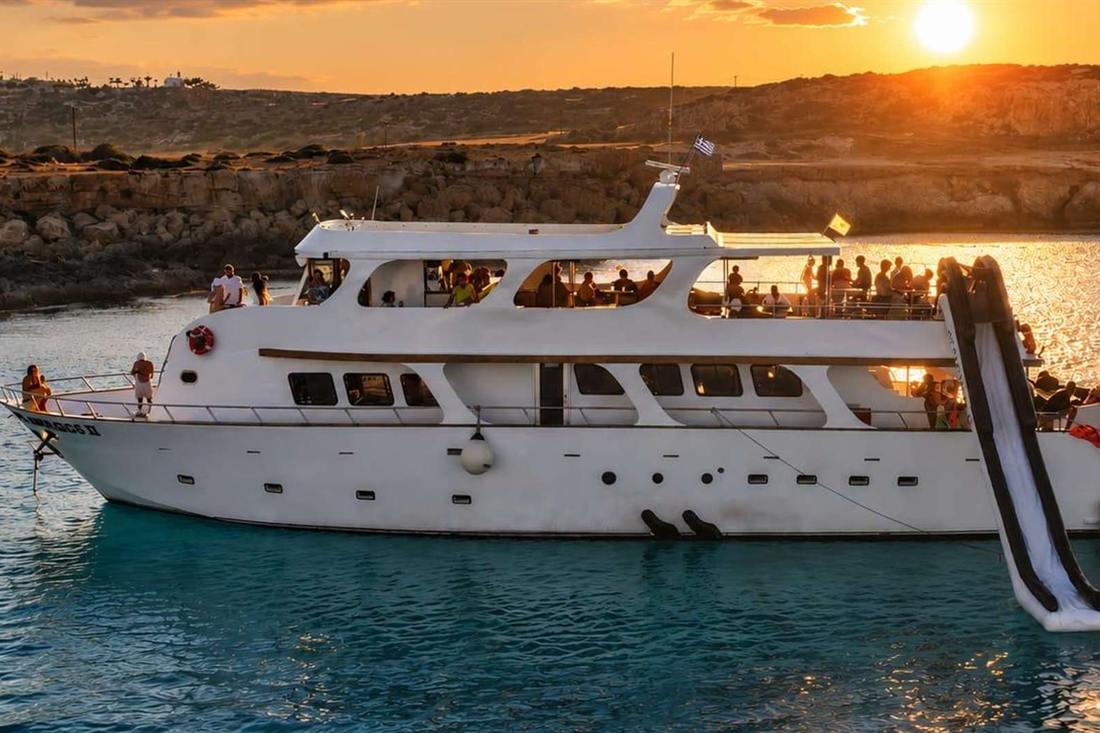 White charter yacht with a large black and white inflatable slide at its stern, anchored in calm water beside a rocky coast under a low orange sun, passengers on the decks