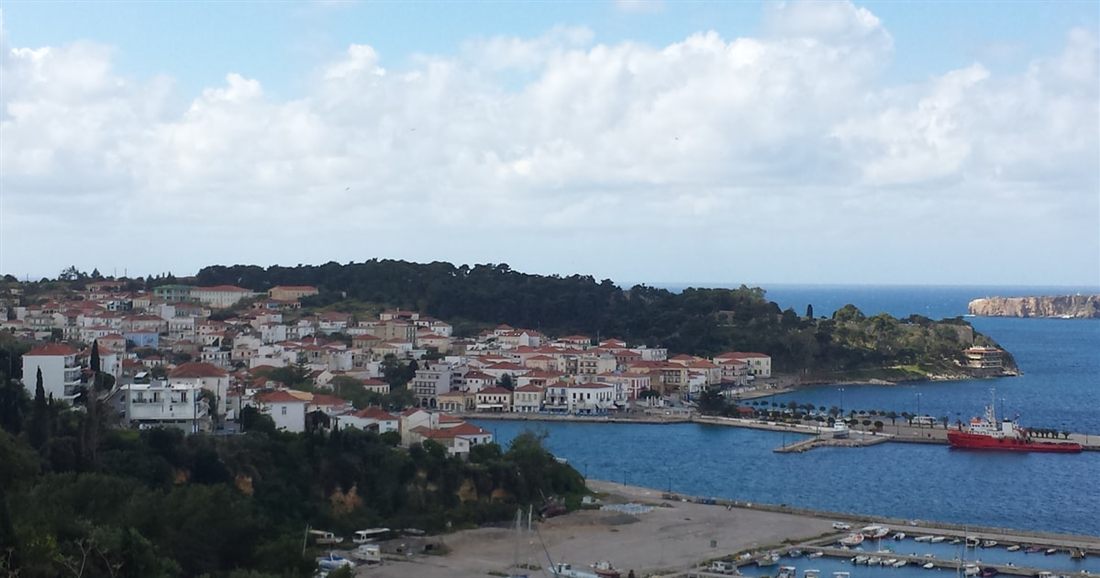 White houses of Pylos above the harbour on Navarino Bay, with a red boat at the quay and wooded hills behind.