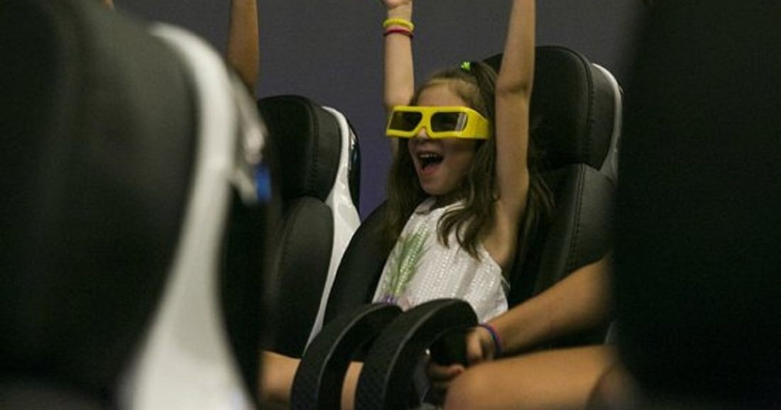 A laughing girl in yellow 3D glasses raises her arms in a moving cinema seat during the Rhodes 9D history film.