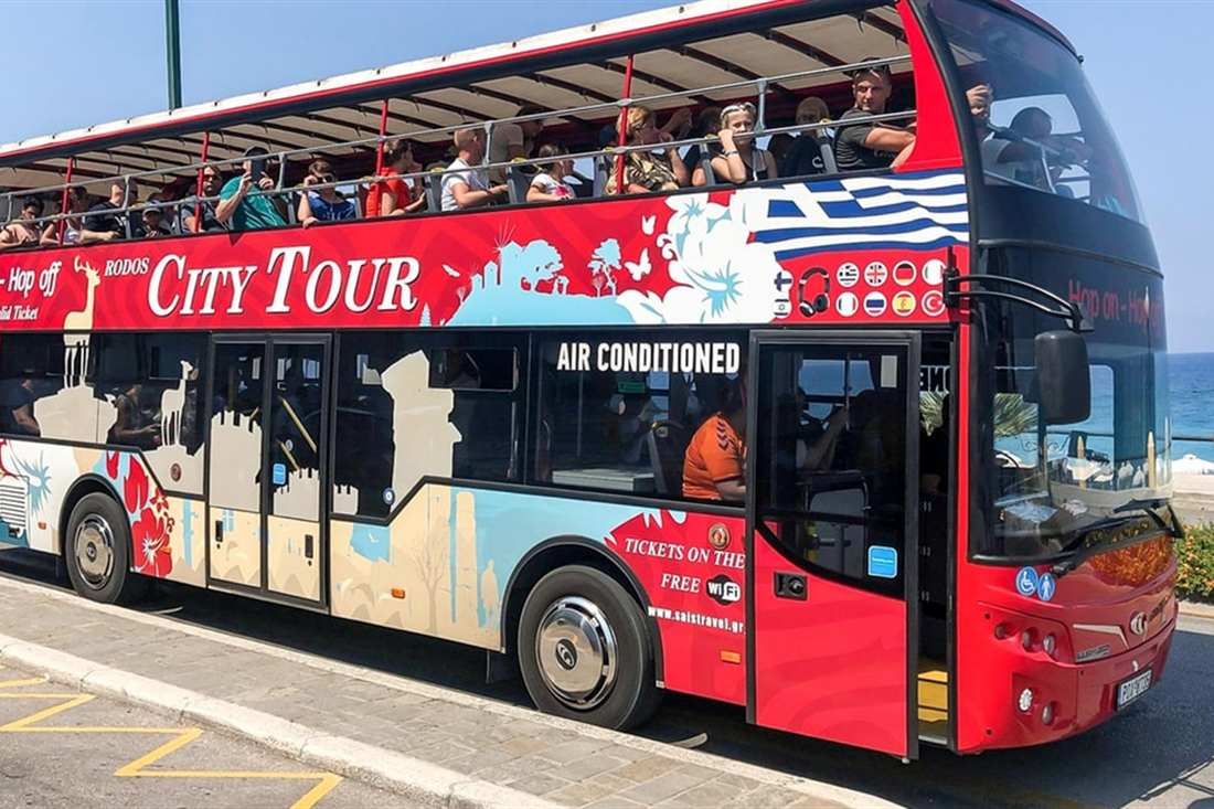A red open-top double-decker City Tour bus with passengers on the upper deck parked by the sea in Rhodes Town, used for Rhodes hop on hop off.