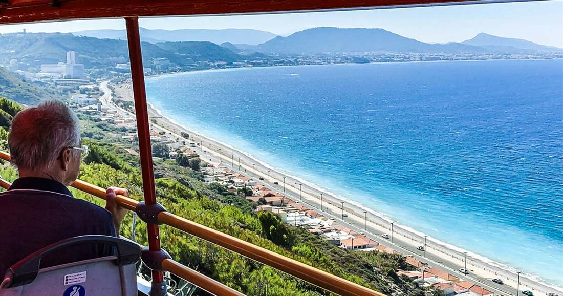 A passenger on the open top deck of a sightseeing bus looks over a long curved beach and blue sea on Rhodes during Rhodes hop on hop off.