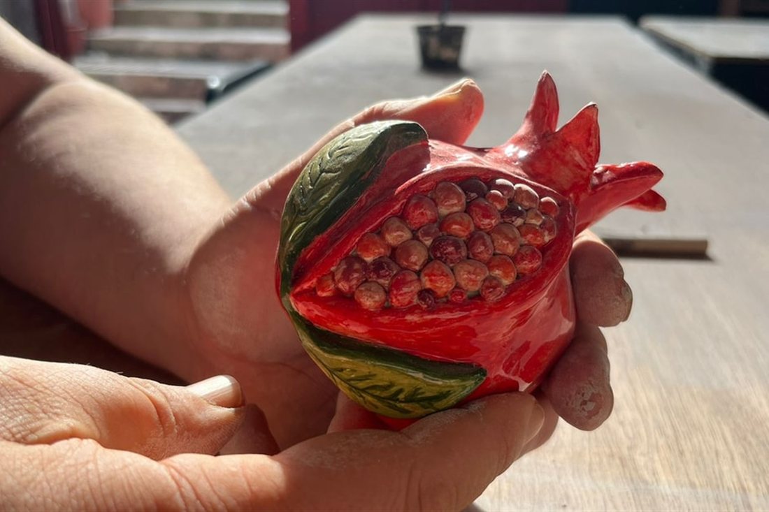 Hands holding a finished red ceramic pomegranate with painted seeds and leaves, made in a Rhodes pottery class.