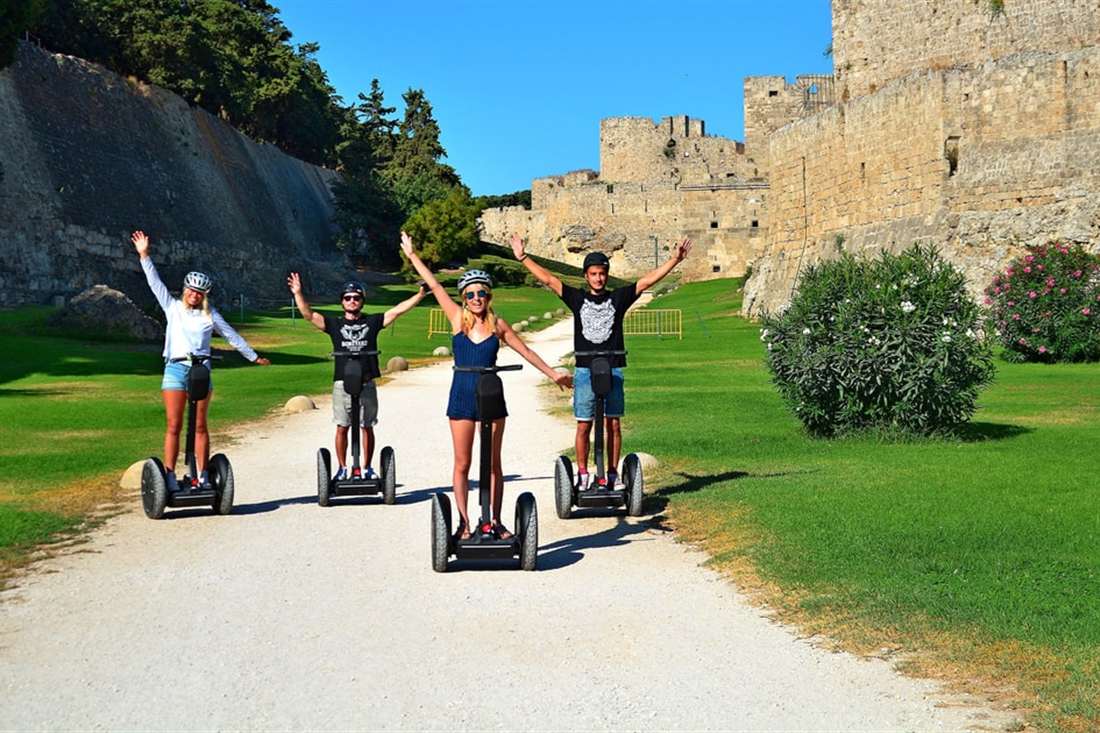 Four riders on segways with their arms raised on a path beside the medieval walls of Rhodes on a Rhodes segway tour.