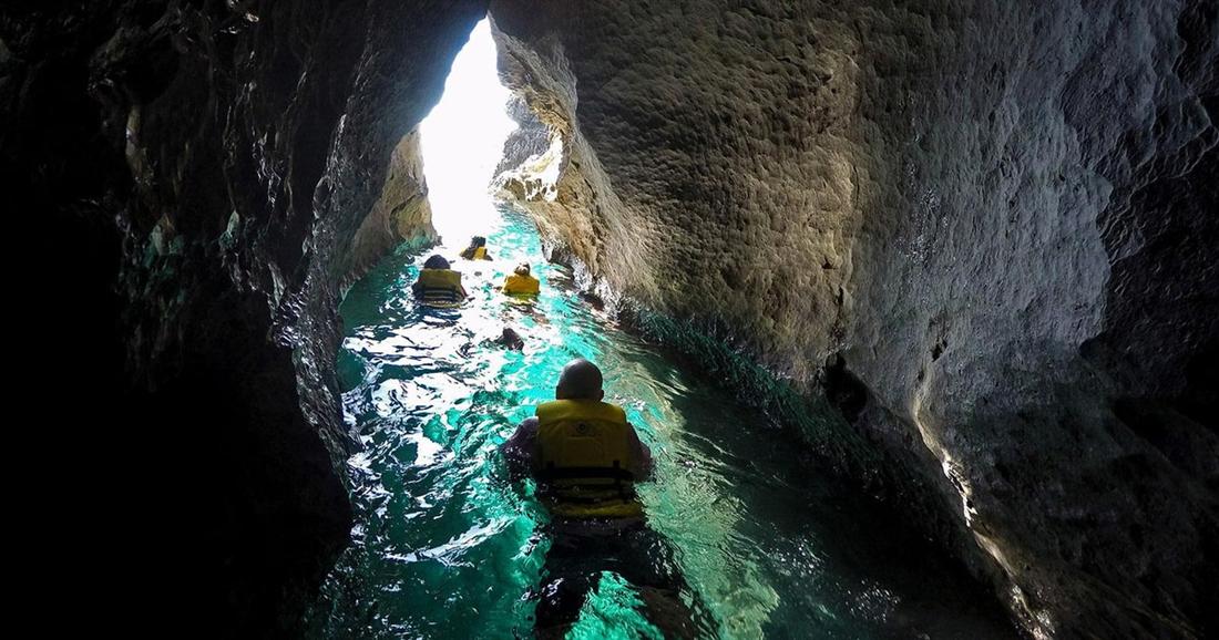 A paddler in a yellow life jacket rides a paddleboard through a narrow sea cave with green water and rock walls, on Rhodes stand up paddle.