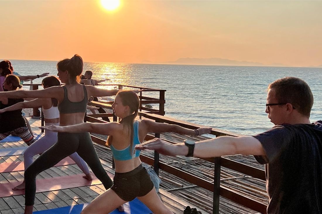 A group holding a warrior pose on a wooden deck by the sea as the sun sets, during a Rhodes yoga class at Kremasti.