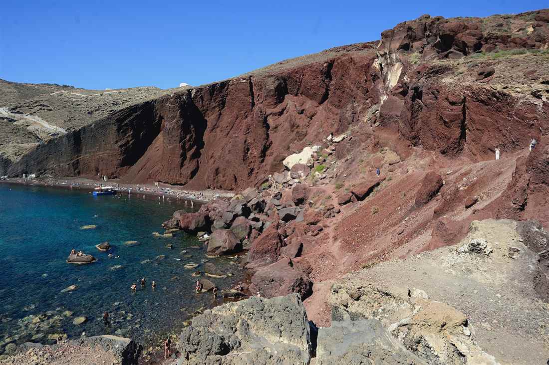 The red volcanic cliffs above the dark sand and clear water of Red Beach near Akrotiri, a stop on a Santorini bus tour.
