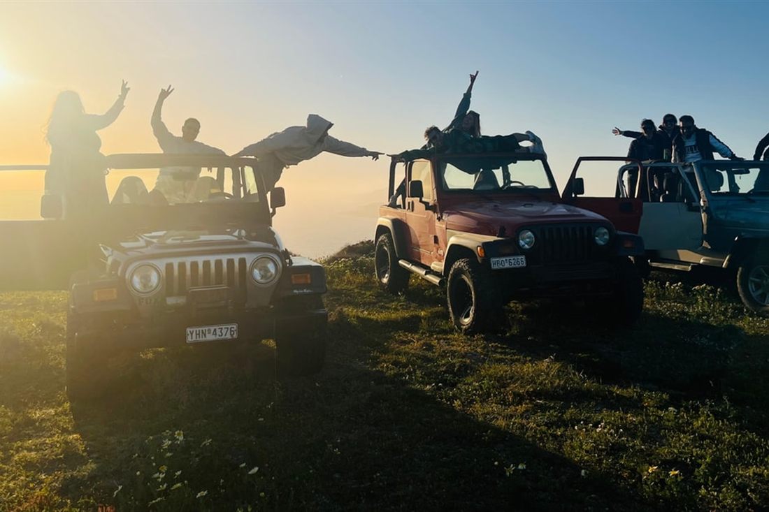 People stand and wave on top of open Wrangler jeeps parked on a grassy hill at sunset, on a Santorini jeep tour.