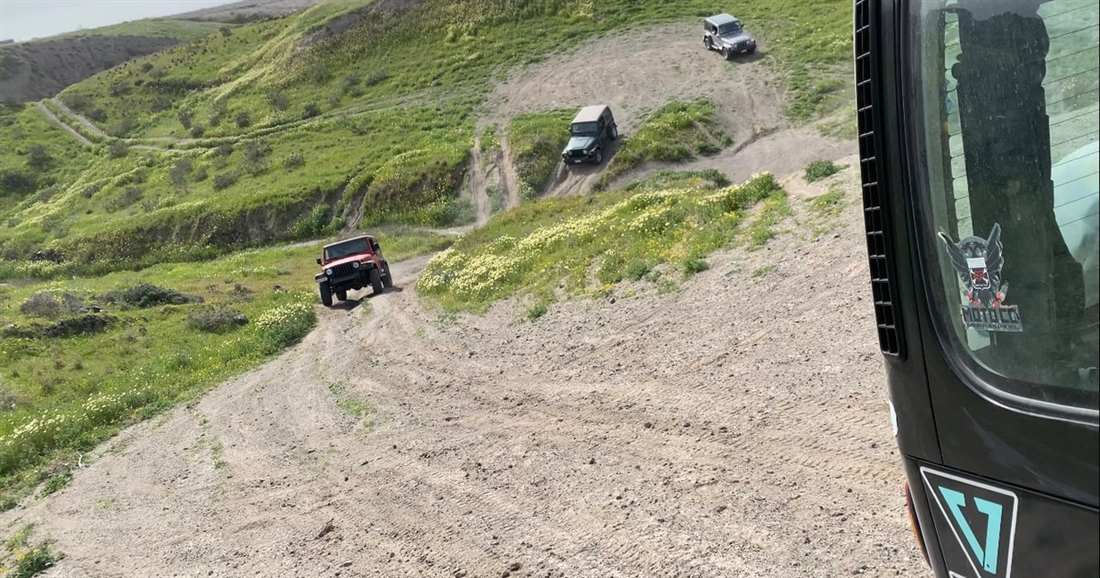 Jeeps drive down a dirt track across green hillsides, seen from inside another jeep on a Santorini jeep tour.