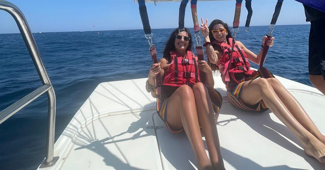 Two smiling riders in red life jackets sit strapped into the harness on the deck of a parasailing boat at sea, before a Santorini parasailing flight.