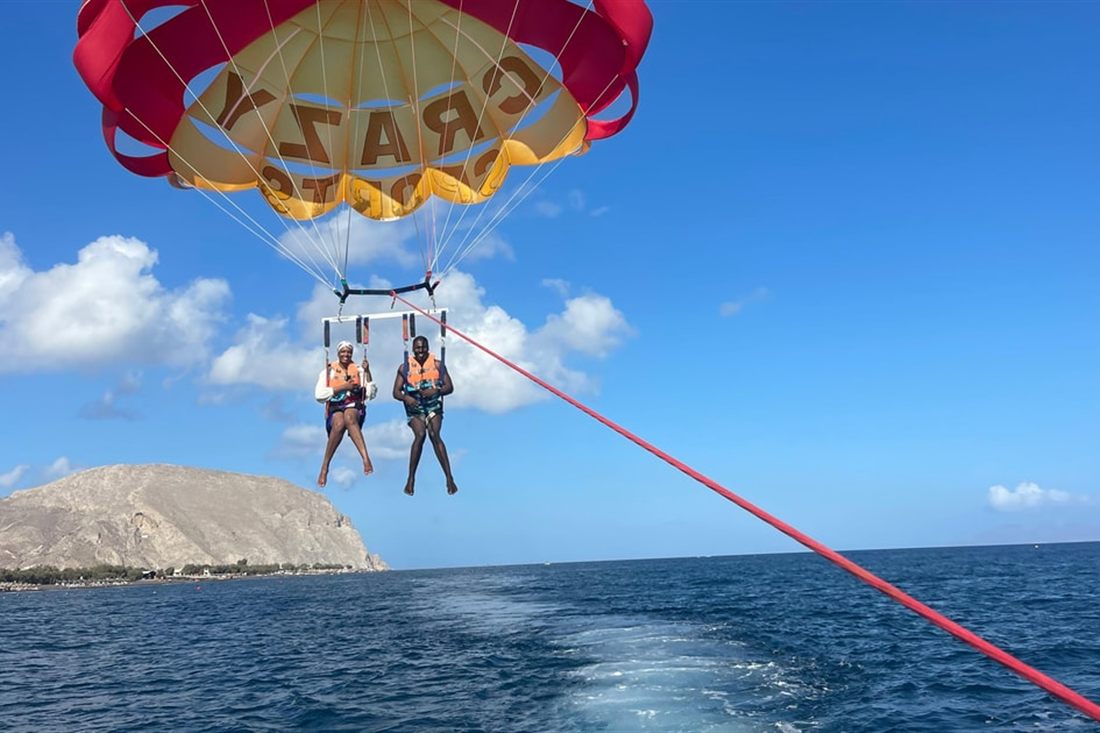 Two riders hang below a red and yellow parasail over the blue sea, with the boat wake and a rocky headland behind them, on Santorini parasailing.