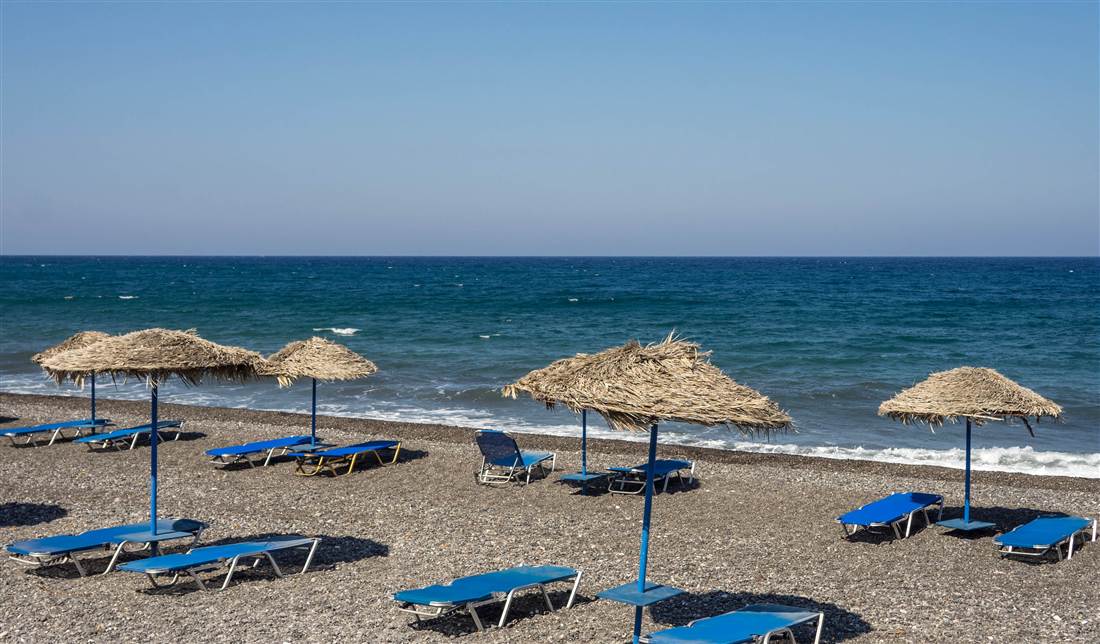 Straw umbrellas and blue sunbeds on the black sand of Kamari beach, where a Santorini spa therapy is held by the sea.