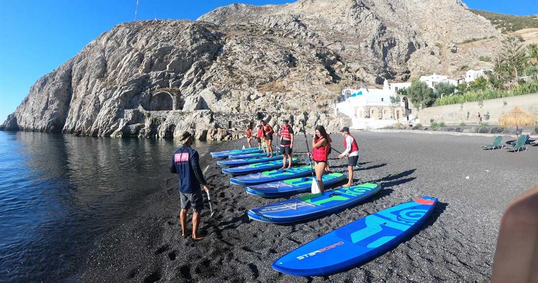 SUP boards lined up on a black sand beach below a cliff while a guide briefs the group before a Santorini SUP tour.