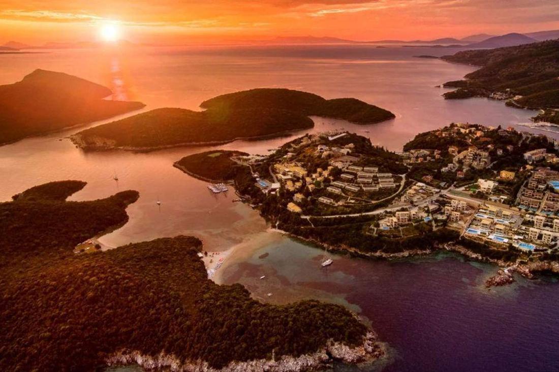 Aerial view at sunset of forested islets, a coastal village with swimming pools and a sandy cove, with the sun low over the sea