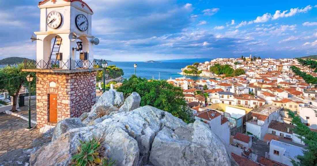 A stone church clock tower stands on rocks above the red roofs of Skiathos town, with the sea behind.