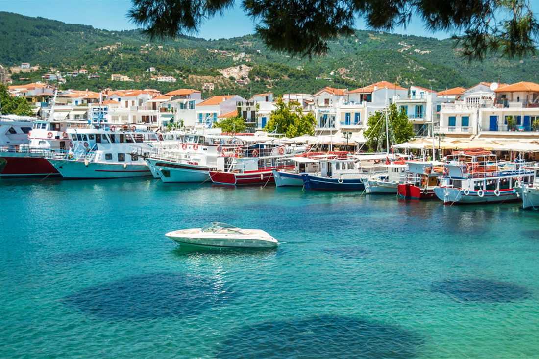 Small boats moor in clear green water in front of white houses at the harbour of Skiathos town.