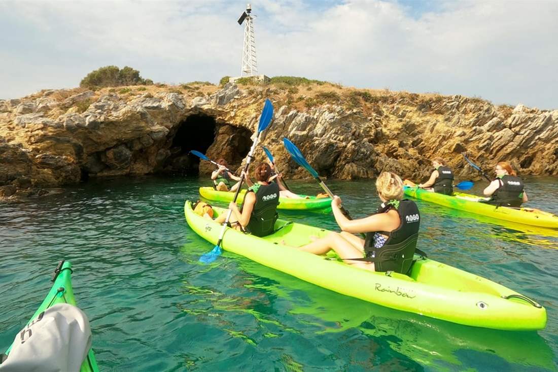 Kayakers in green sit-on-top kayaks paddling past a rocky islet with a sea cave and a small beacon on a Skiathos kayak tour.