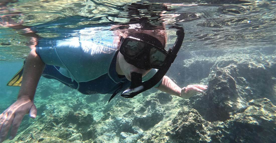 A snorkeler in a mask and wetsuit swimming over a rocky reef in clear shallow water