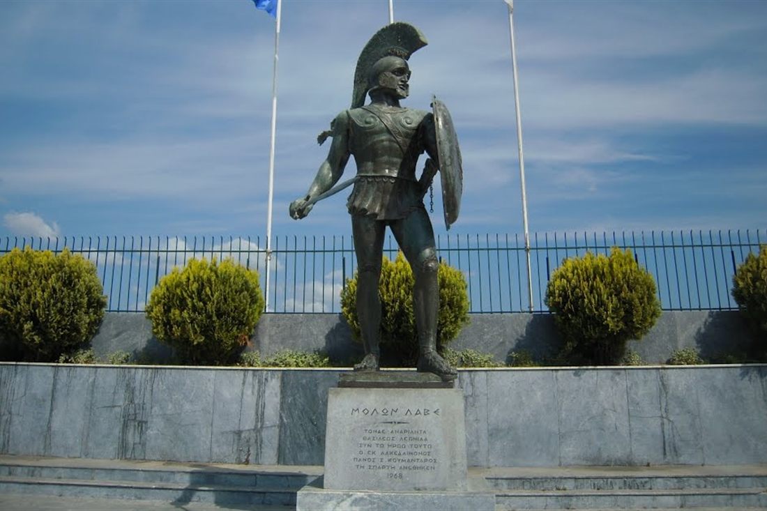 The bronze statue of Leonidas with a helmet, spear and round shield on a marble base in Sparta, under a blue sky.