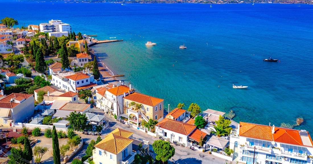 Red-roofed houses of Spetses along a blue coast, seen from above, with small boats on the water.