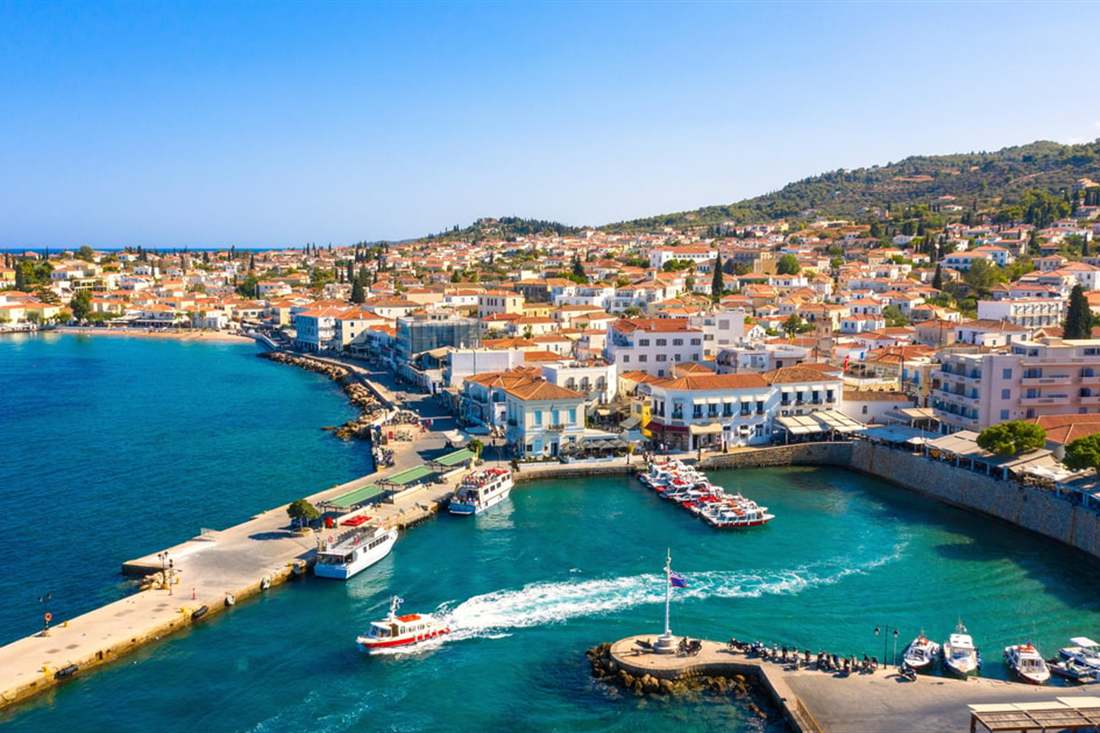Aerial view of the harbour of Spetses town, with a long quay, moored boats, a boat wake and houses on the hill behind.