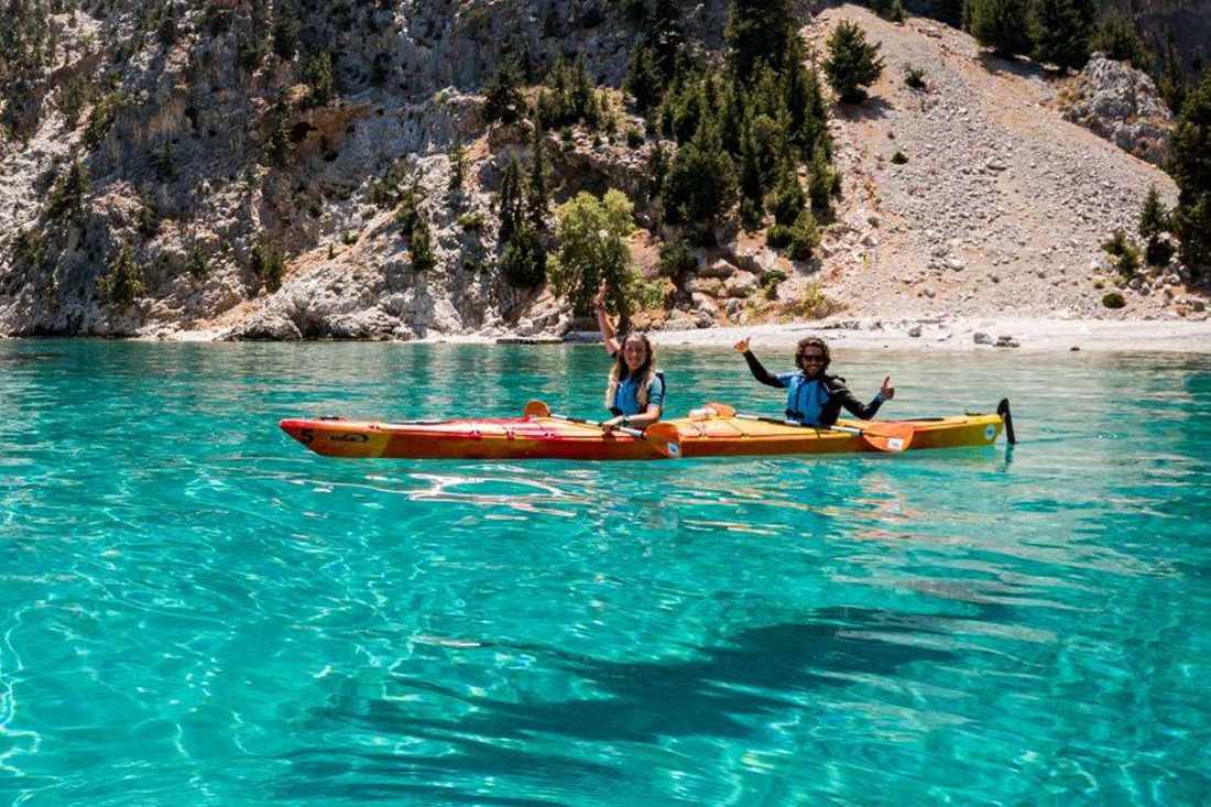 Two people paddling a double sea kayak on turquoise water beside a rocky hillside and a pebble beach