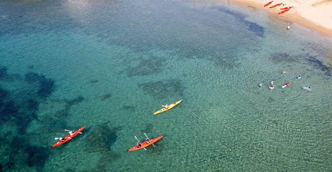 Red and yellow sea kayaks paddling over clear turquoise water near a sandy cove with kayaks beached