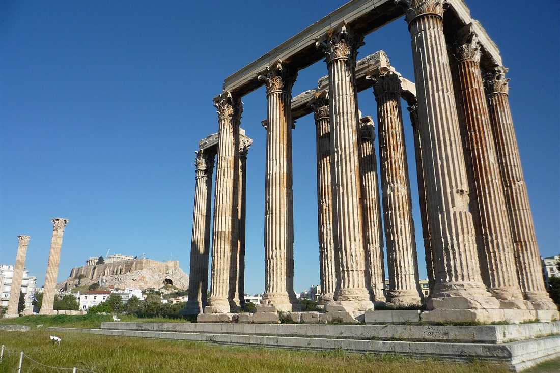 The Corinthian columns of the Temple of Olympian Zeus under a deep blue sky, with the Acropolis on its rock in the background.
