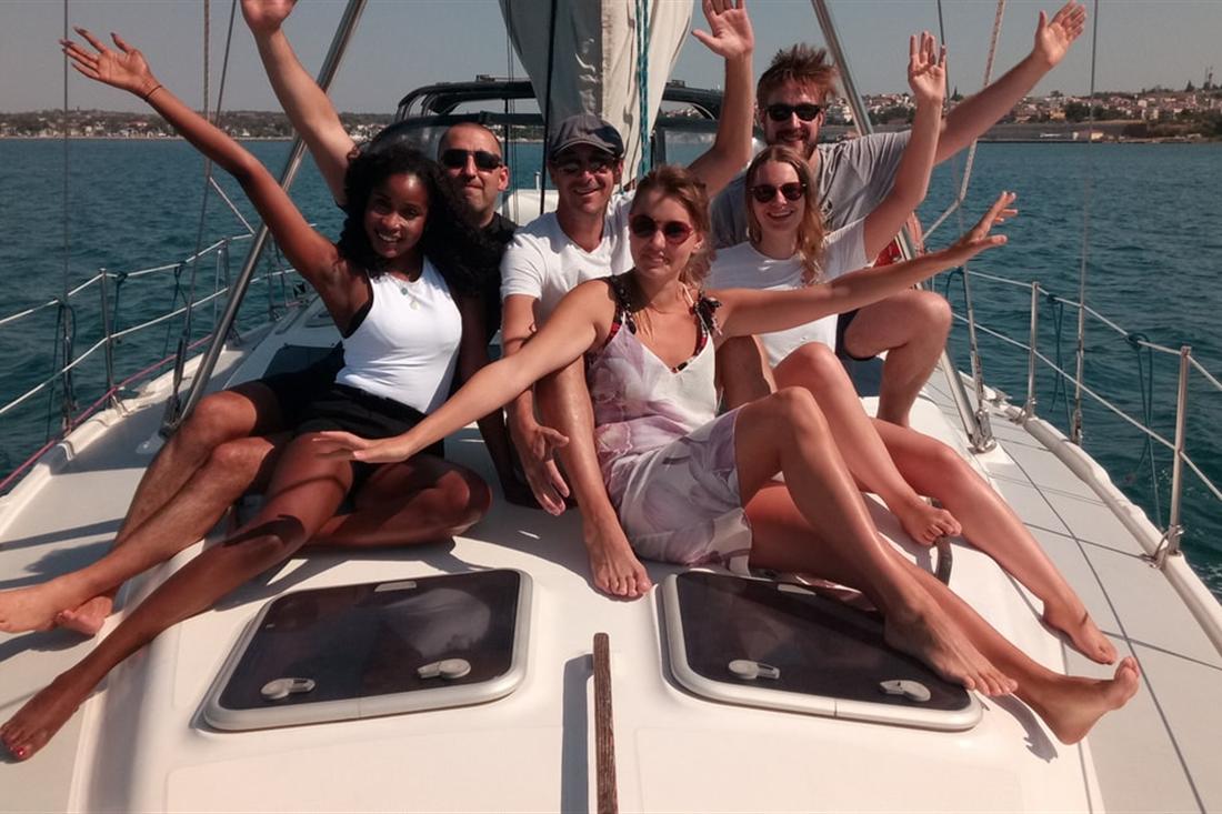 A group of smiling guests sitting on the bow of a white sailing yacht on the open sea, with a low shoreline behind them, on a Thessaloniki boat tour.