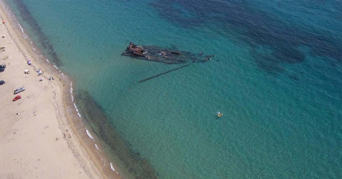An aerial view of a half-sunken rusty shipwreck in clear green water off a long sandy beach at Epanomi, the stop on the Shipwreck Bay Thessaloniki boat tour.