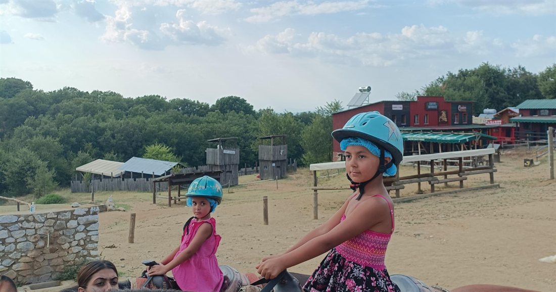 Two children in riding helmets on horseback in a sandy riding ring at a farm, on Thessaloniki horse riding.