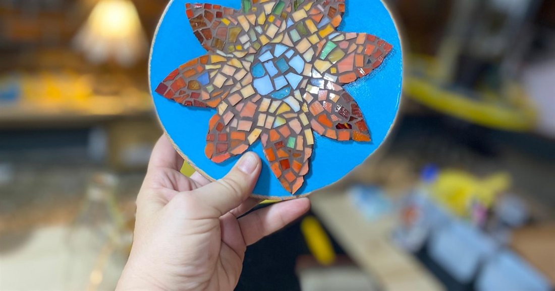 A finished glass mosaic of an orange and blue flower on a round board, made in a Thessaloniki mosaic workshop.