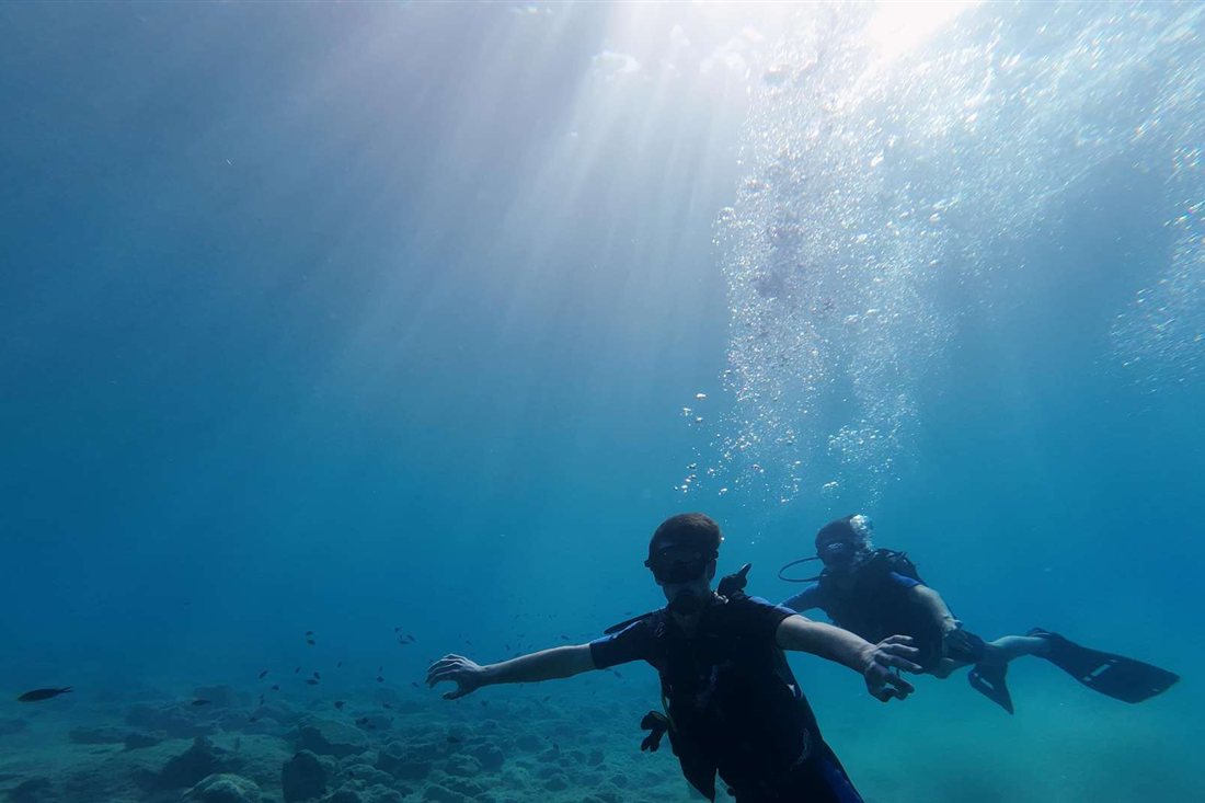Two scuba divers swim in clear blue water with sun rays above on a dive from Tolo