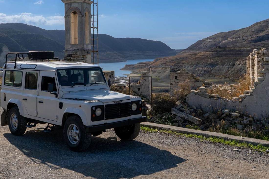 White Land Rover Defender parked on a gravel road beside ruined stone walls and a stone bell tower, with a reservoir and brown mountain slopes behind under a blue sky