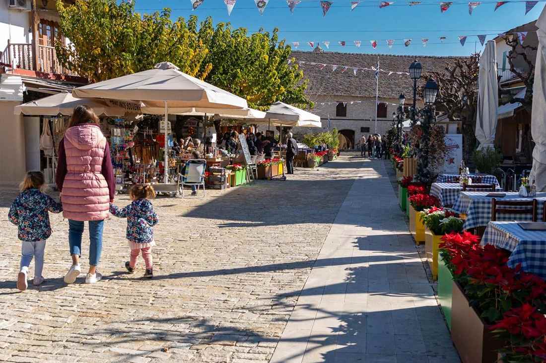 Cobbled village square in the mountains with market stalls under umbrellas, cafe tables with blue checked cloths, red flowers and trees, and a woman walking with two small children