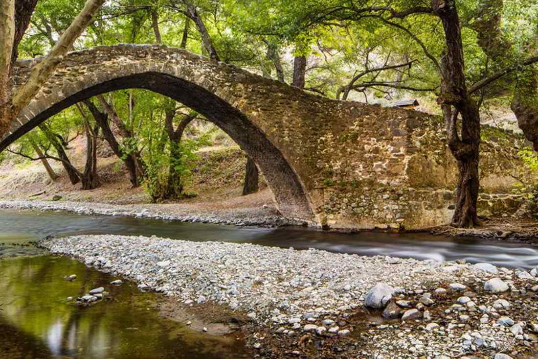 Old single-arch stone bridge over a shallow river with a pebble bank and plane trees in the Troodos mountains of Cyprus