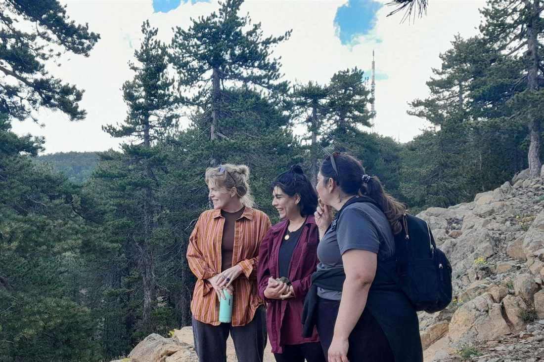 Three hikers smiling on a rocky mountain trail among tall black pines, with a radio mast on the summit behind them
