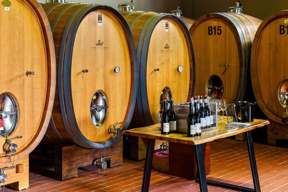 Large oak wine barrels and a tasting table with bottles and glasses in a Tyrnavos winery