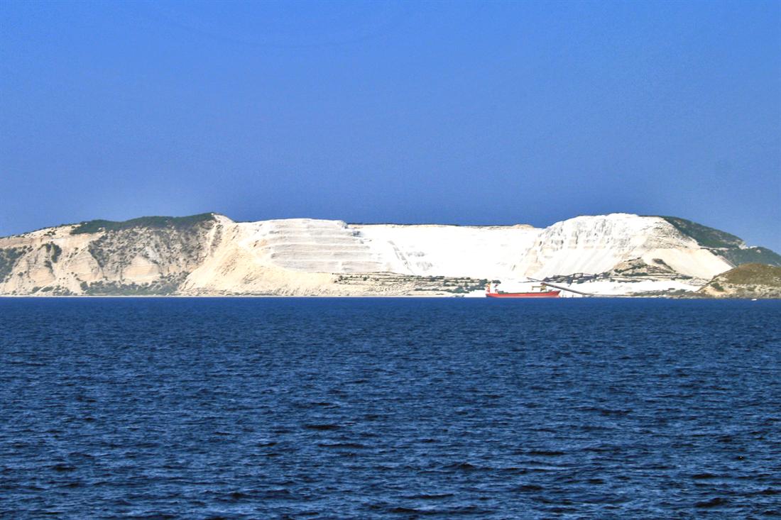 White pumice quarry slopes on Yali island rising above dark blue sea, with a red cargo ship at the shore