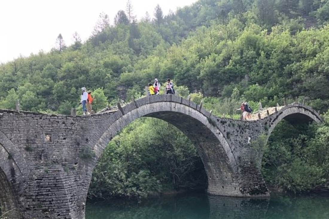 Visitors walk over the three-arched Kalogeriko stone bridge above a green river pool in the forest of Zagori.
