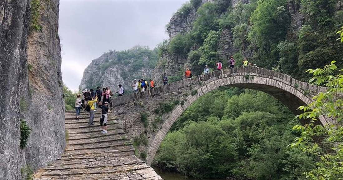 Walkers cross a high single-arch stone bridge with stepped paving between a rock wall and green forest in Zagori.