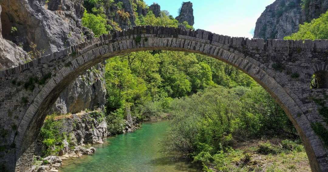 A single-arch stone bridge over a turquoise river in a green gorge in Zagori.