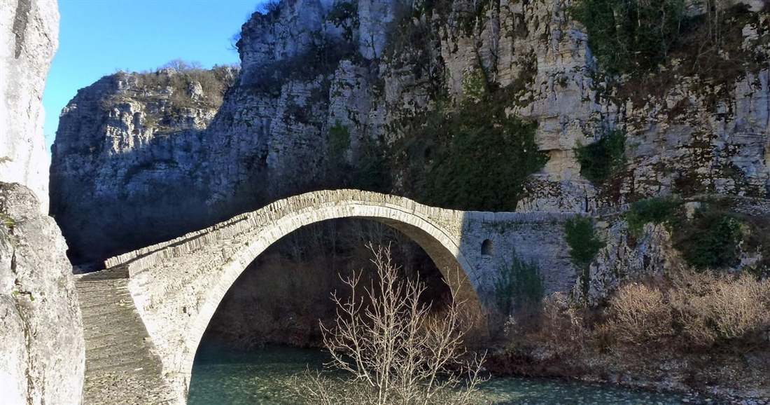 A single high stone arch bridge crosses a clear river below grey limestone cliffs in Zagori.