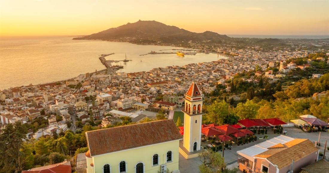 Zakynthos Town and its harbour at sunset seen from the hill above, with a church bell tower in front, a viewpoint on a Zakynthos e-bike tour.