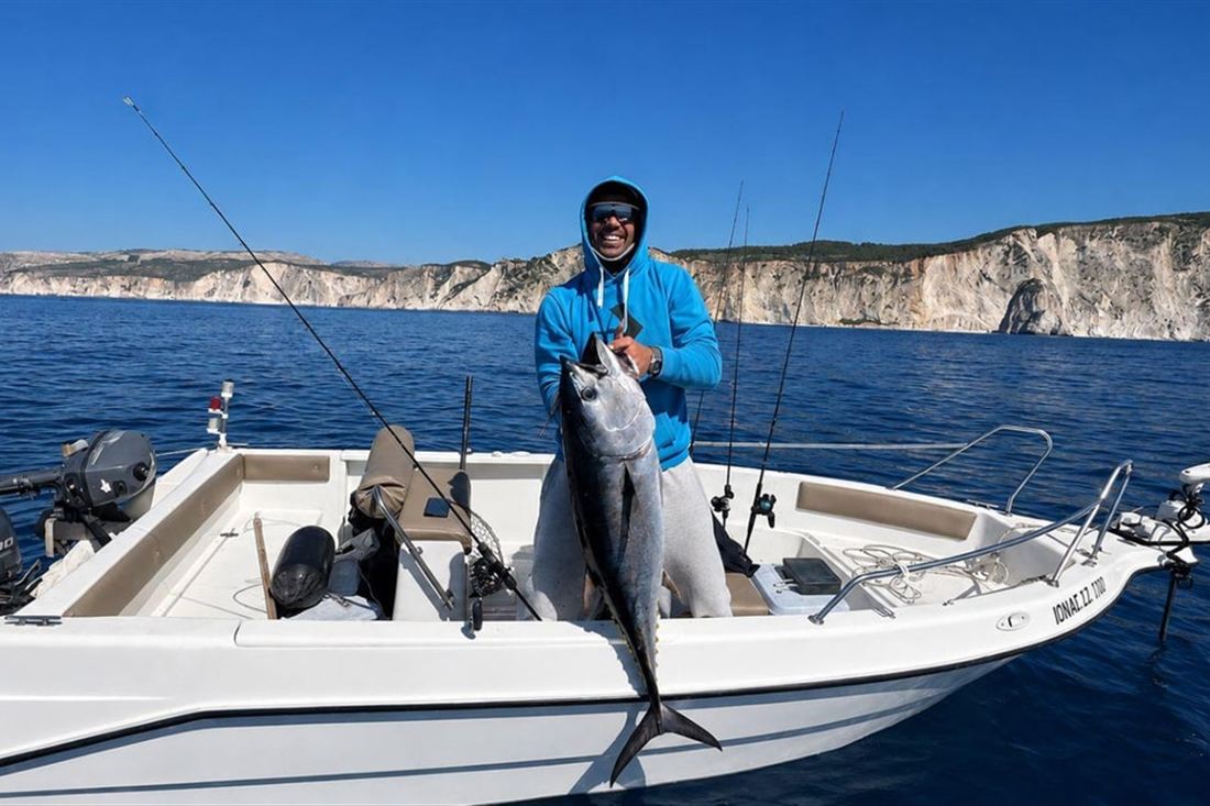 An angler on a small fishing boat holding a tuna, with the white cliffs of the Zakynthos coast behind, on a Zakynthos fishing trip.
